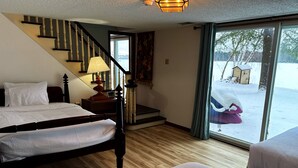 A hotel room with a wooden floor, a bed with white bedding, a nightstand with a lamp, a staircase, and a view of a snowy landscape through a large window. - Foster's Lakeside - Sandy Shore with Snowmobile Access & Year-Round Fun (Tomahawk)