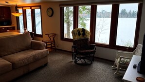 A living room with a sofa, a rocking chair, a dining table, and a view of a lake through large windows. - Foster's Lakeside - Sandy Shore with Snowmobile Access & Year-Round Fun (Tomahawk)