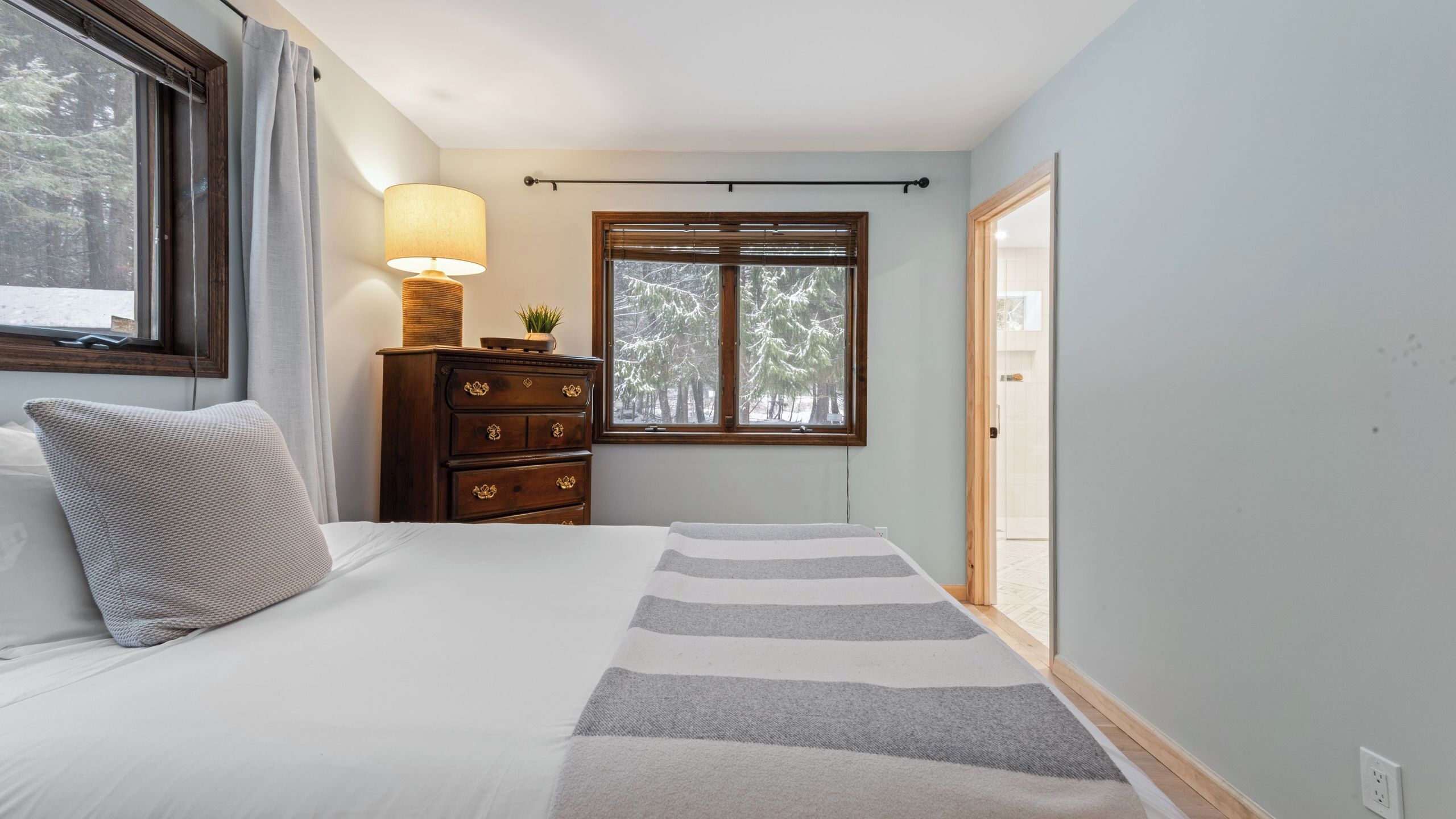 Une chambre à coucher comprenant un lit, une commode, une lampe, une fenêtre donnant sur des arbres et une porte menant à l’extérieur.