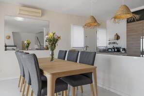 A dining area with a wooden table, grey upholstered chairs, a vase of flowers, a mirror, and a kitchen area with a stove and utensils. - 3306 (Mijas Costa)