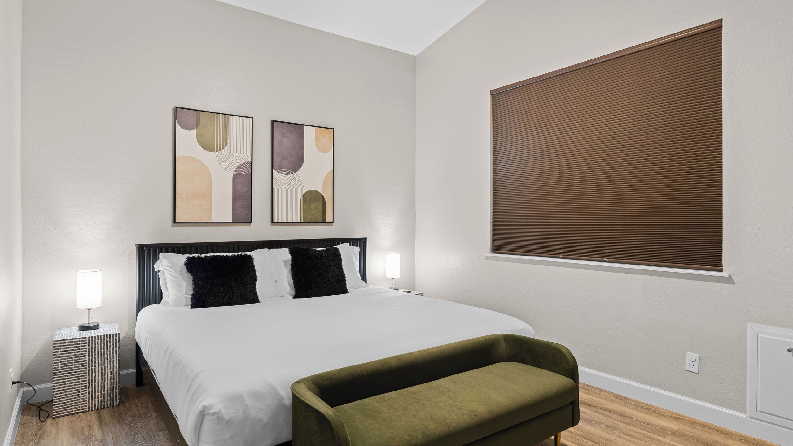 Una camera da letto moderna con un letto grande, due opere d'arte appese al muro, una finestra con tende e un comodino con una lampada.