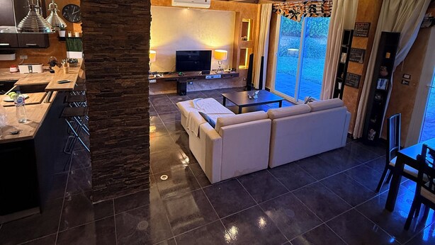 A modern living room with a large white sofa, a kitchen area with a stone accent wall, and a dining area with a view of the outdoors.
