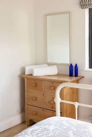 Un dormitorio con un aparador de madera, una cama blanca, un espejo y dos botellas azules sobre el aparador.