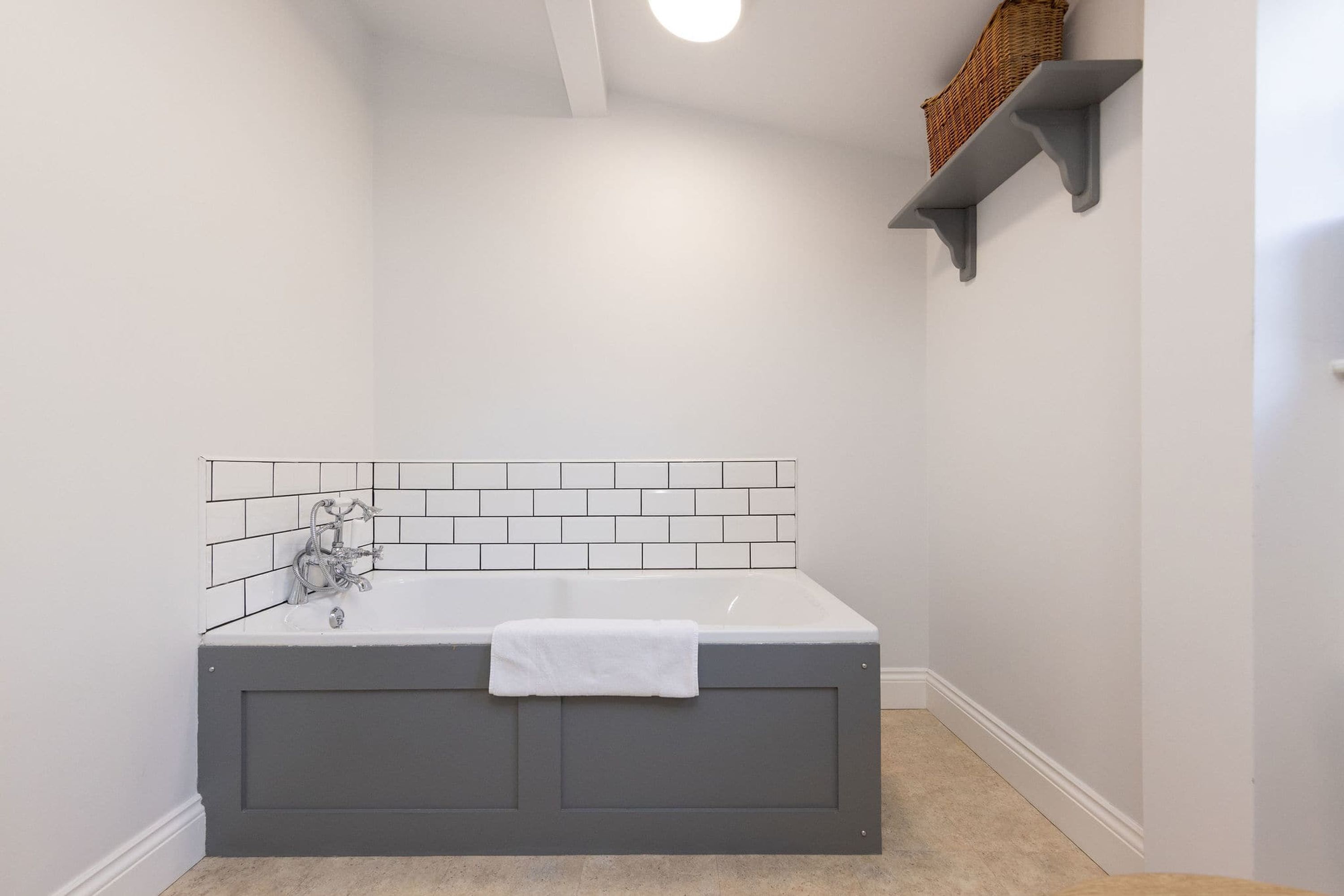 Une salle de bain avec une baignoire, des murs recouverts de carreaux blancs, une serviette blanche et une étagère avec un panier.