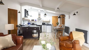 Un salon moderne avec un canapé en cuir marron, une table basse et un coin repas comprenant une table et des chaises en bois. On aperçoit au fond une cuisine avec des meubles sombres et un îlot central.