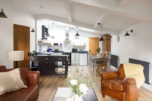 A modern living room with a brown leather sofa, a coffee table, and a dining area with a wooden table and chairs. A kitchen with dark cabinets and a central island is visible in the background. - Sidlesham Hideaway Logburner Garden (Chichester)