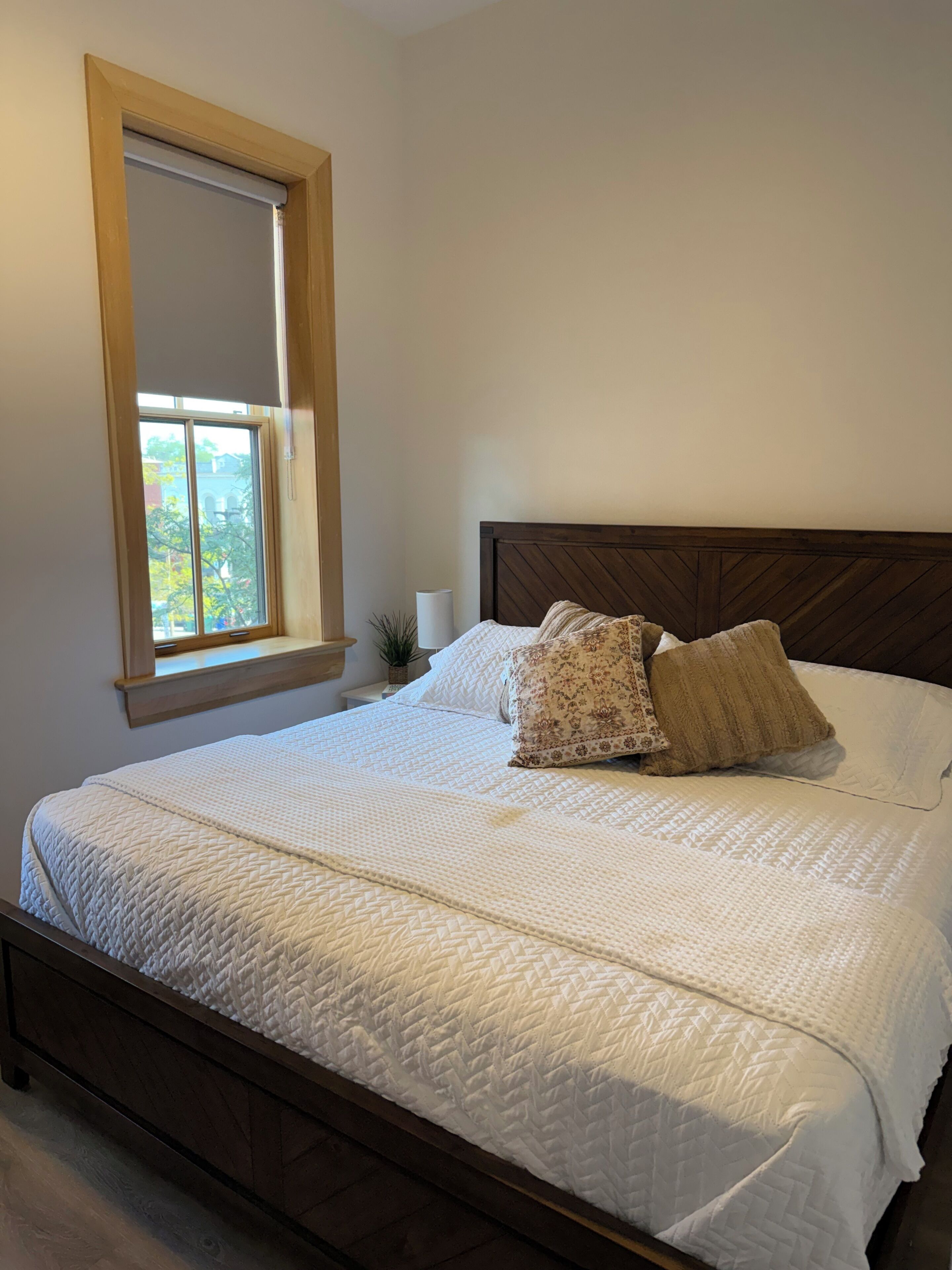 Een slaapkamer met een houten bedframe, wit bedgoed, een raam met uitzicht en een kleine plant op het nachtkastje.