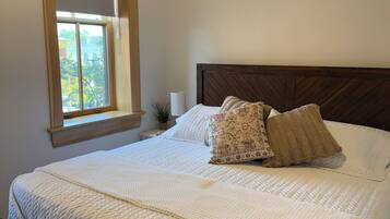 Een slaapkamer met een houten bedframe, wit bedgoed, een raam met uitzicht en een kleine plant op het nachtkastje.