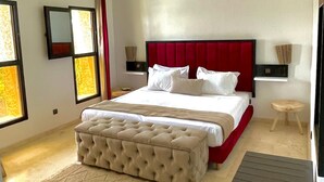 A modern bedroom with a large bed, a tufted headboard, a bedside table, a small stool, and a large window. - Luxury villa heated pool (Marrakech)