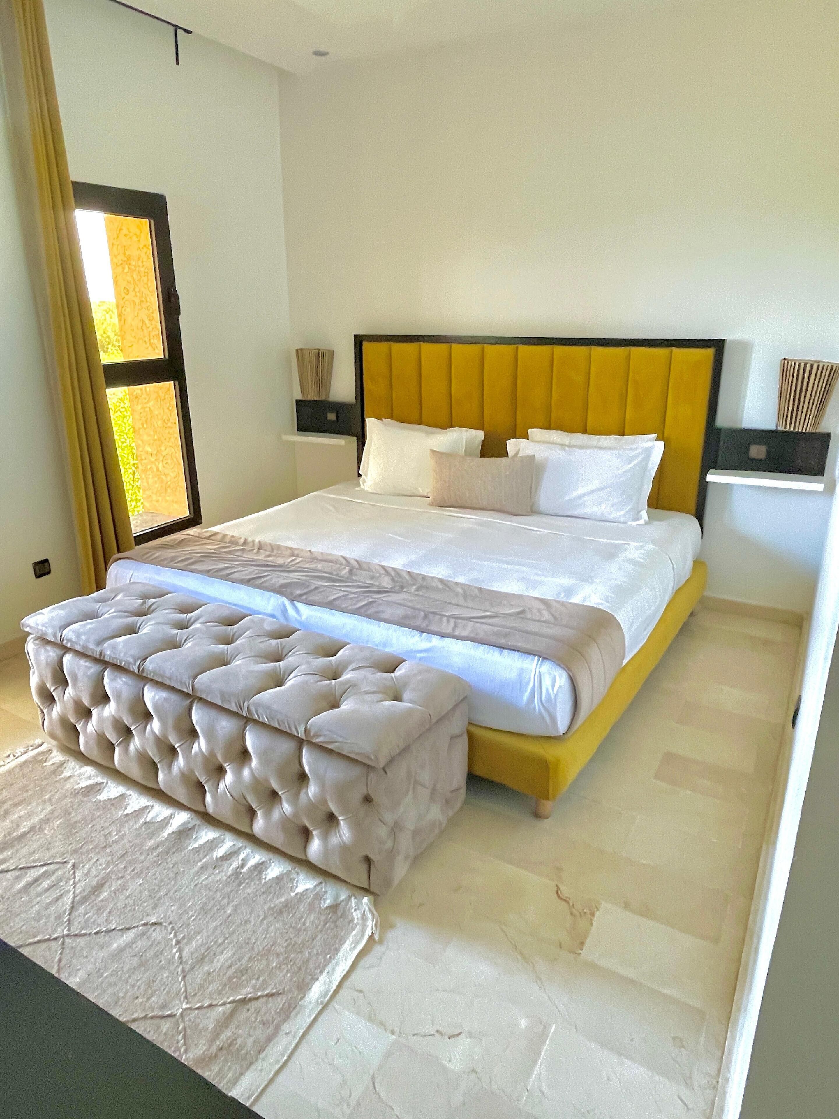 Une chambre à coucher moderne avec un grand lit, un pouf capitonné et une table de chevet avec une lampe.