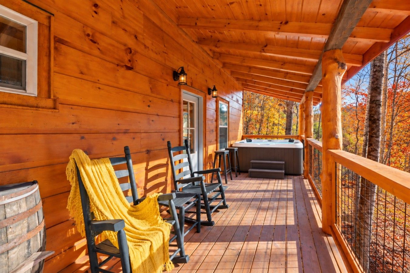 A wooden deck with a hot tub, chairs, and a covered area with a view of autumn trees.