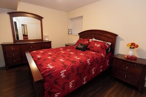 Quarto com cama de madeira, mesa de cabeceira, cômoda, espelho e janela com persianas.