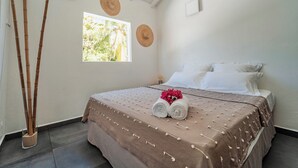 Un lit bien fait, recouvert d’une couverture marron, avec deux serviettes enroulées et un arrangement floral décoratif. Deux chapeaux tressés sont accrochés au mur, il y a une fenêtre donnant sur un paysage verdoyant et une plante de bambou dans un pot.