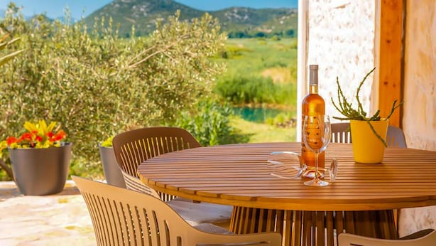 A round wooden table with chairs, a bottle of wine, and a glass on it, set outdoors with a view of greenery and hills in the background.
