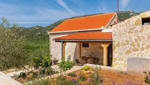 A stone house with a red-tiled roof, a garden with various plants and flowers, and a mountainous backdrop.