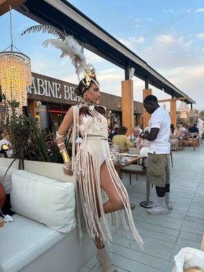 A person in a fringe dress and feathered headpiece sitting on a white couch outdoors, with a restaurant named 'ABINE BEN' in the background. - Villa Acacias – A Peaceful Retreat with a Large Pool in Elviria (Marbella)