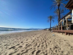 A sandy beach with footprints, palm trees, and a wooden boardwalk with signs. - Villa Acacias – A Peaceful Retreat with a Large Pool in Elviria (Marbella)