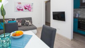 Ein modernes Wohnzimmer mit einem grauen Sofa, einem Esstisch mit Obstschale und einem an der Wand befestigten Fernseher.