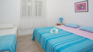 Ein Schlafzimmer mit zwei Betten, einem Fenster mit durchsichtigen Vorhängen, einem Nachttisch mit Lampe und einem gerahmten Bild an der Wand.