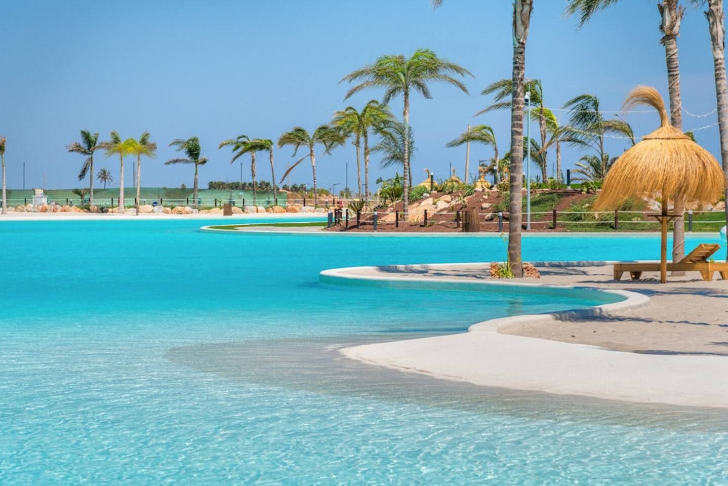 A resort pool with clear turquoise water, surrounded by palm trees and a sandy beach.