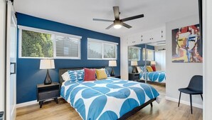 A bedroom with a bed, two bedside tables, a chair, a ceiling fan, and a large window with blinds.