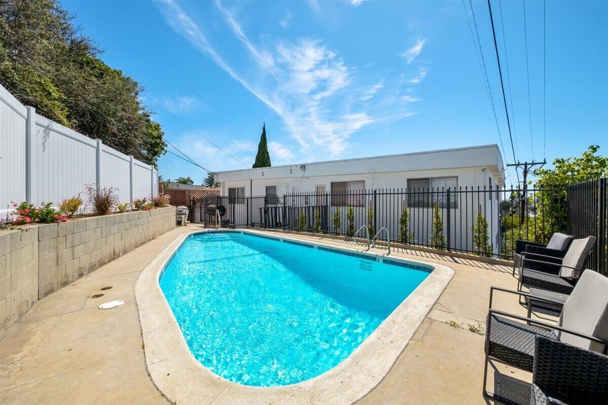 Ein rechteckiger Pool mit Betonrand, umgeben von einer Betonplattform. Auf der Plattform stehen schwarze Metallstühle und ein kleiner Tisch mit einem Stuhl. Im Hintergrund ist ein weißes Gebäude mit schwarzem Zaun zu sehen.