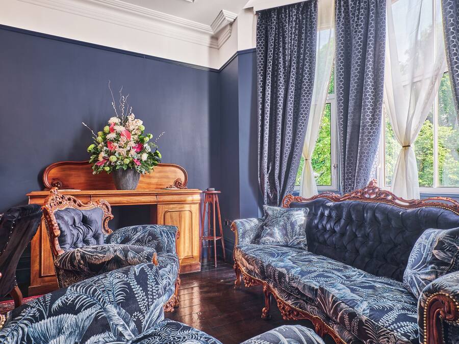 Una habitación con pared azul oscuro, un escritorio de madera con un arreglo floral y un sofá de estampado azul.