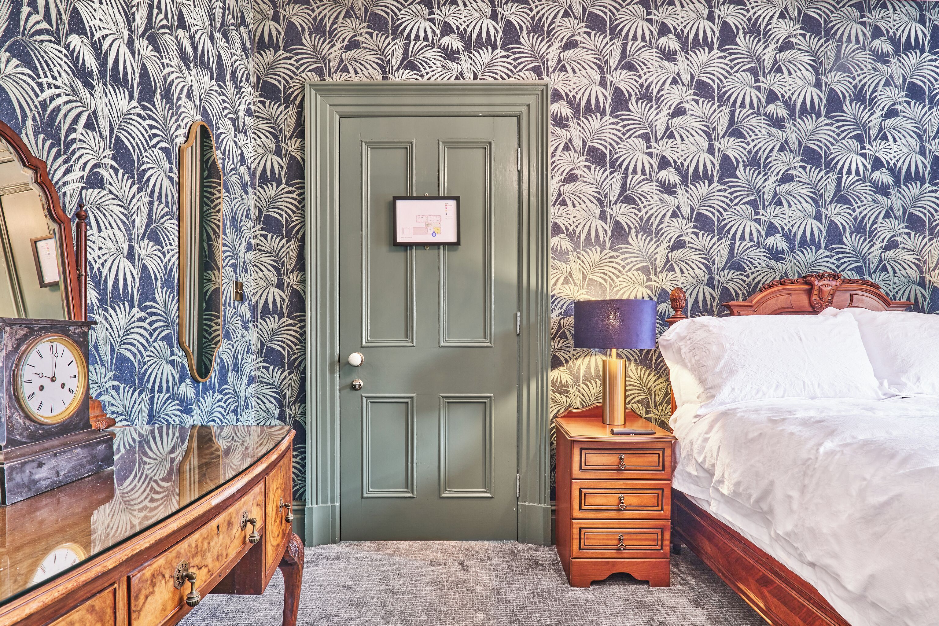A room with a green door, a wooden dresser with a clock, a bedside table with a lamp, and a bed with white bedding.