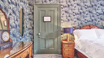 Una habitación con puerta verde, cómoda de madera con reloj, mesita de noche con lámpara y cama con ropa de cama blanca.