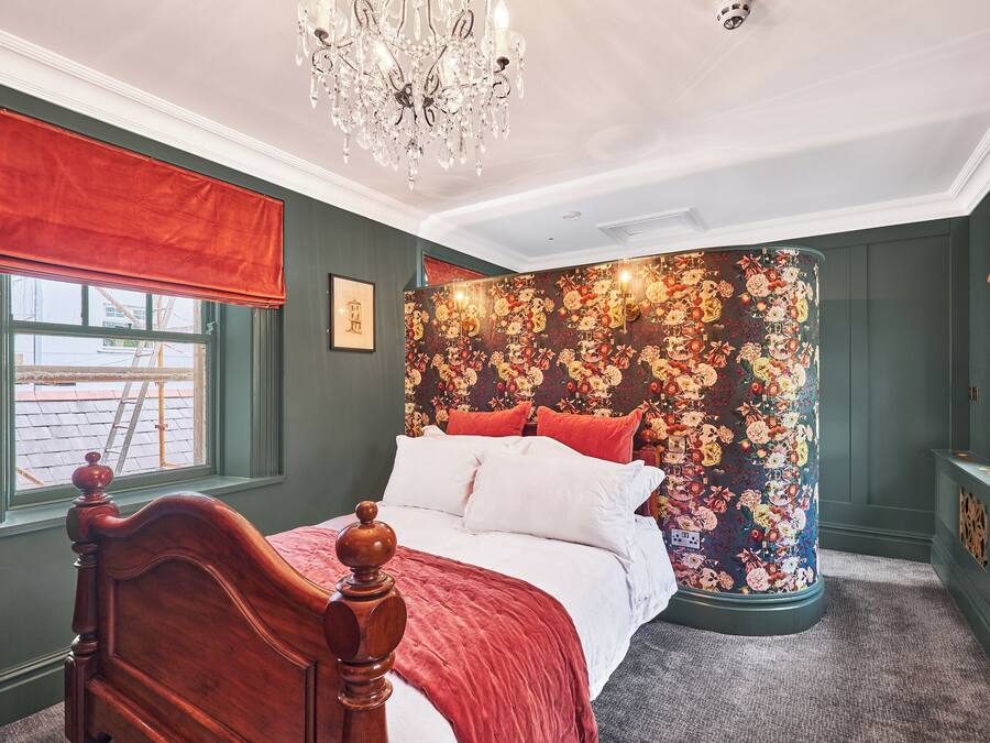Un dormitorio con cama de madera, colcha roja y blanca, pared decorativa con papel tapiz floral y un candelabro.