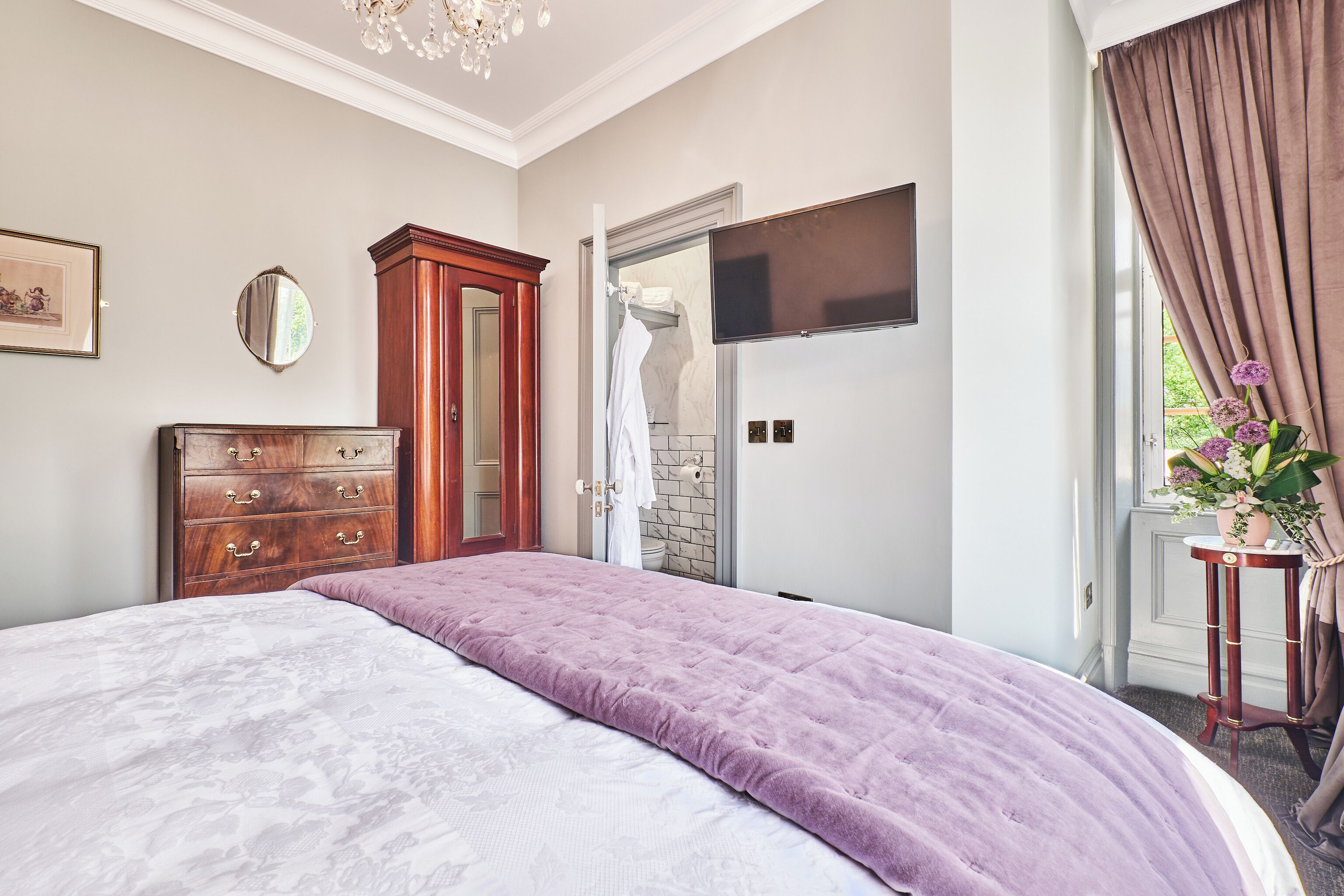A bedroom with a large bed, a wooden dresser, a television on the wall, and a window with curtains.