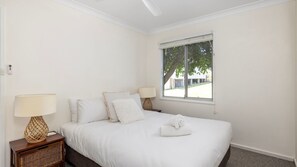 A bedroom with a bed, two bedside lamps, a window, and a view of a house. - Karingal Beach Cottage  Fingal Bay (Fingal Bay)