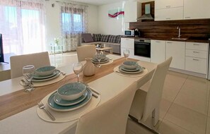 Un coin repas avec une table en bois dressée pour quatre personnes, des chaises blanches et une cuisine en arrière-plan.