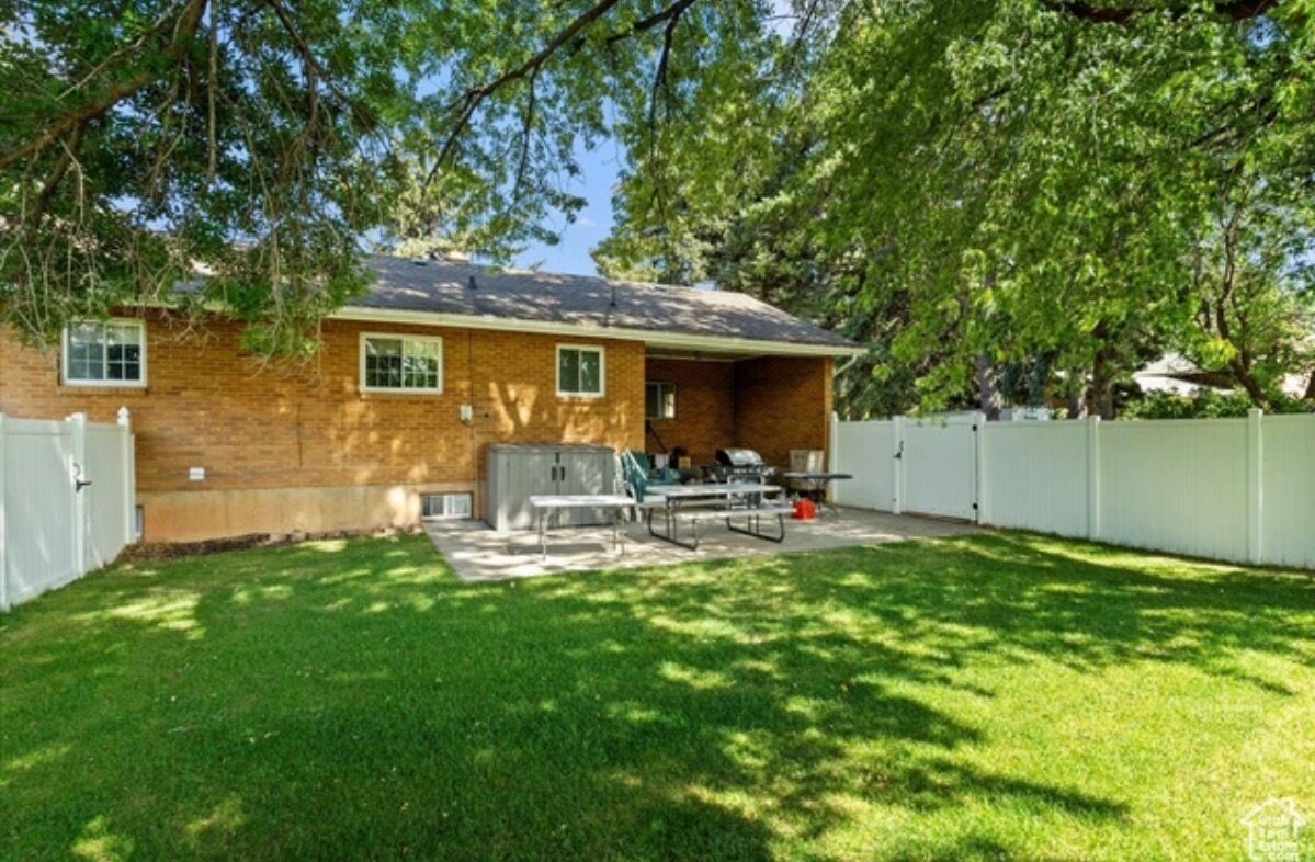 Una casa de un piso de ladrillo con patio trasero cercado, un patio con mobiliario exterior y un árbol grande que da sombra.