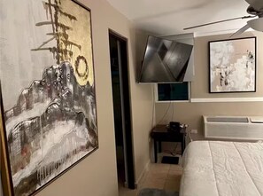Ein Hotelzimmer mit großem abstrakten Gemälde, einem flachbildfernseher an der Wand, einem Bett mit weißer Bettdecke und einer Klimaanlage.