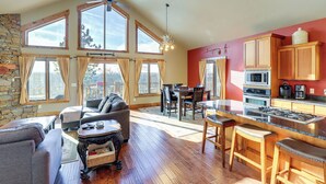 A spacious living area with a fireplace, a dining table, and a kitchen with wooden cabinets and a large window overlooking a scenic view. - 1 Mi to Terry Peak! Black Hills Cabin Hideaway (Lead)