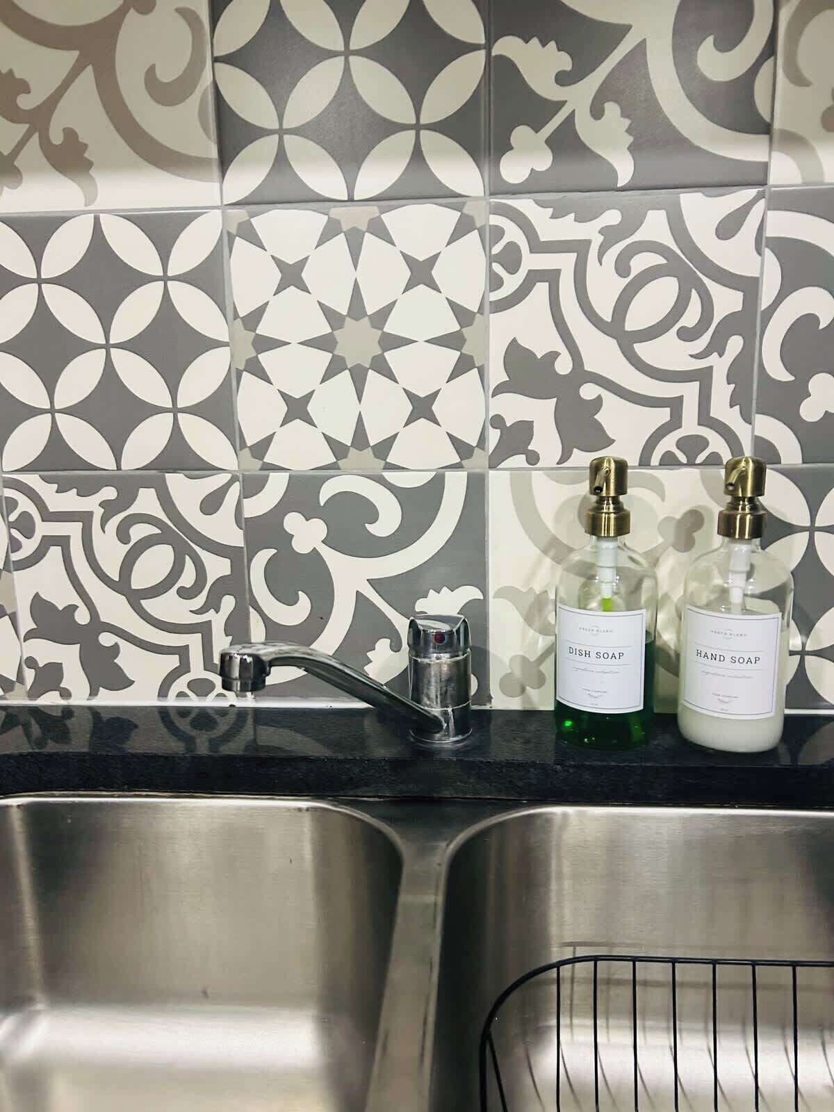 Una cucina con un lavello e un rubinetto moderno, due flaconi di sapone, uno con l'etichetta 'detersivo per piatti' e uno per 'sapone mani', e uno schienale decorativo con motivo geometrico.