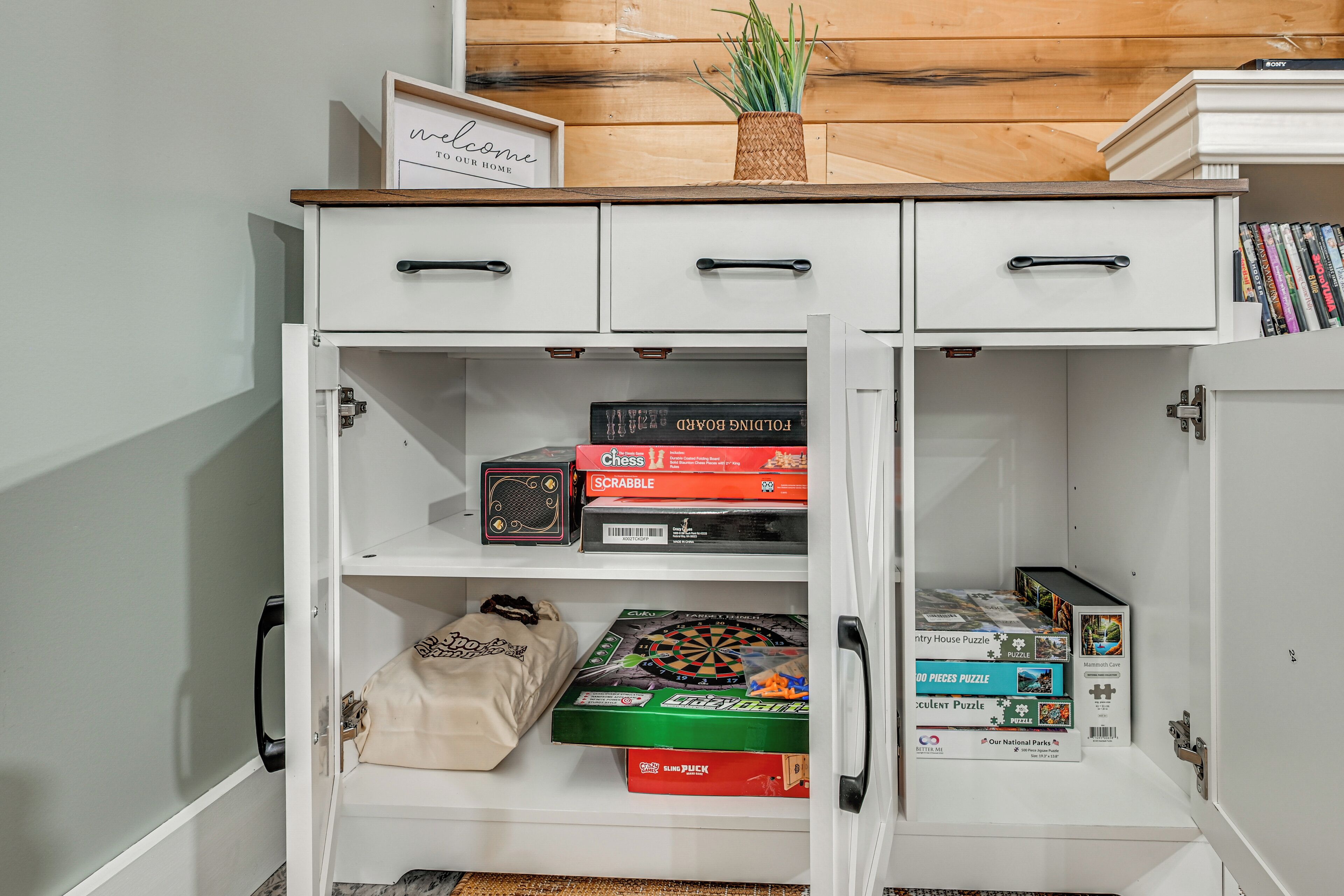 Un mueble blanco con puertas abiertas que muestra juegos de mesa, un juego de ajedrez y un cartel de bienvenida.
