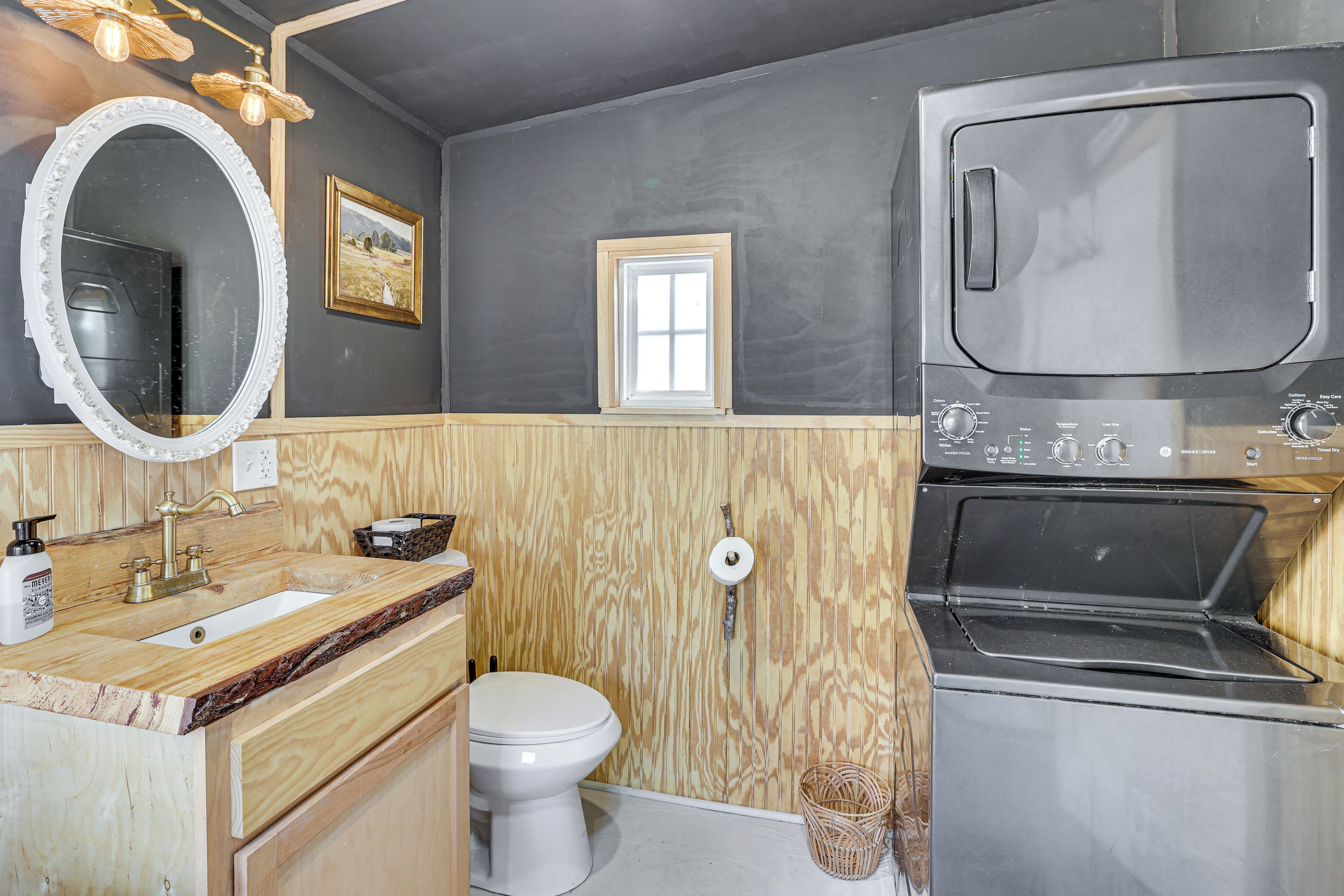 A compact bathroom with a wooden vanity, a white sink, a large mirror, a toilet, and a built-in oven.