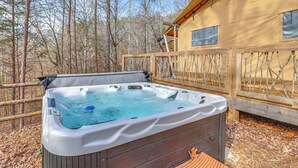 Un jacuzzi en un área boscosa con una terraza de madera y una cabaña al fondo.