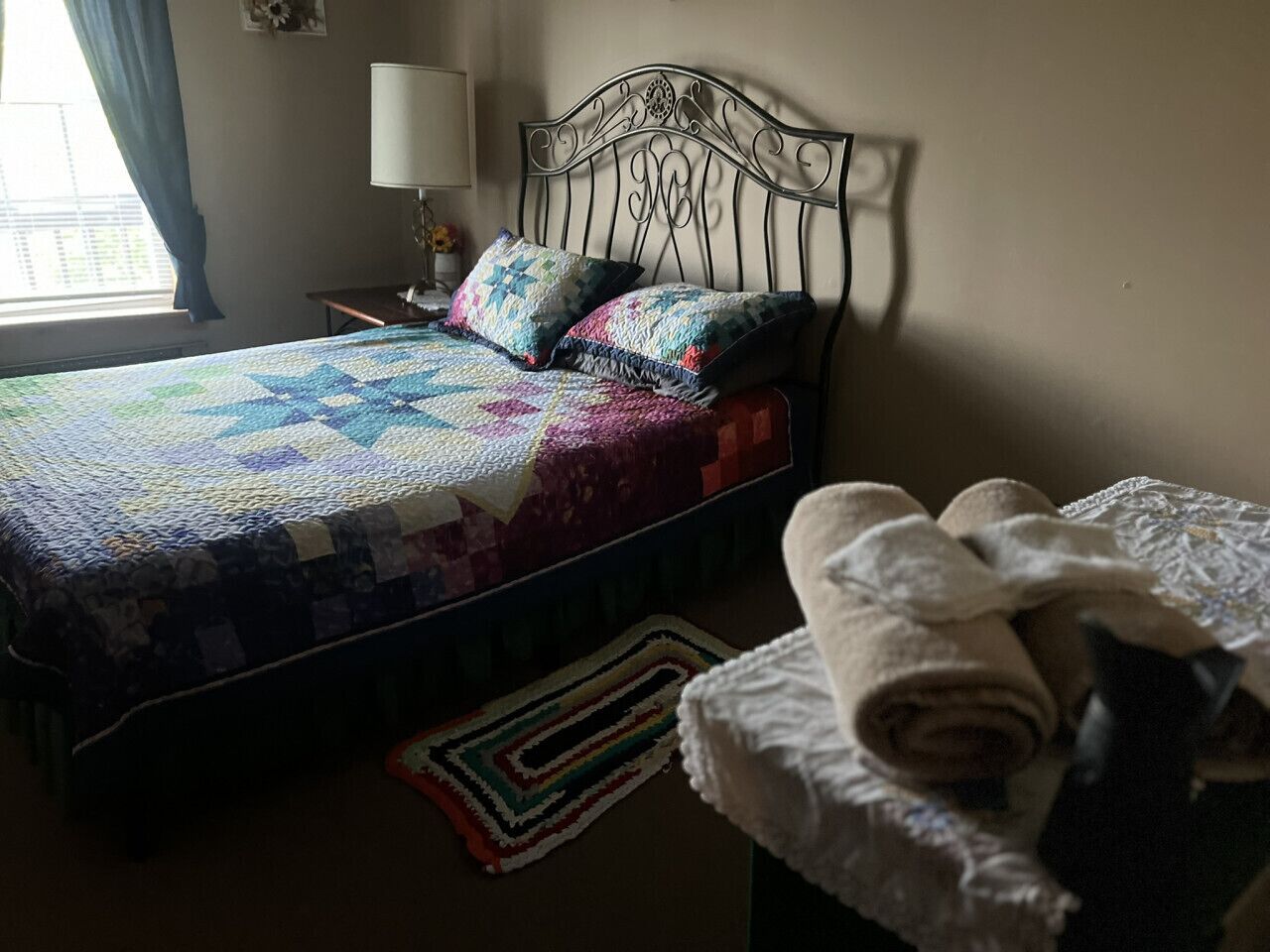 Un lit avec une couverture colorée et des oreillers, une lampe de chevet, une petite table avec une lampe et une serviette enroulée sur une chaise.