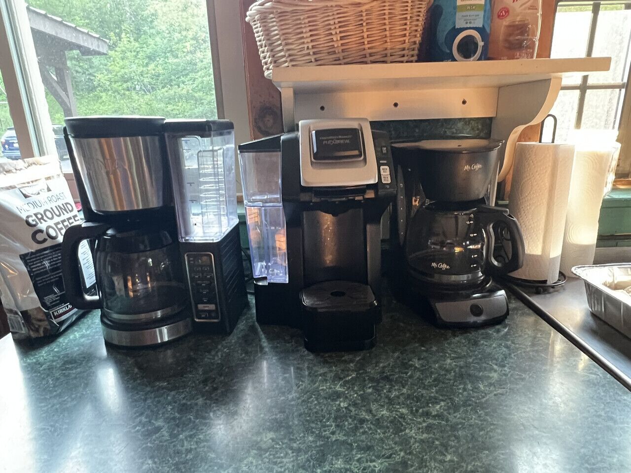 Eine Küchenarbeitsplatte mit verschiedenen Kaffeemaschinen und einem Korb auf der Fensterbank.