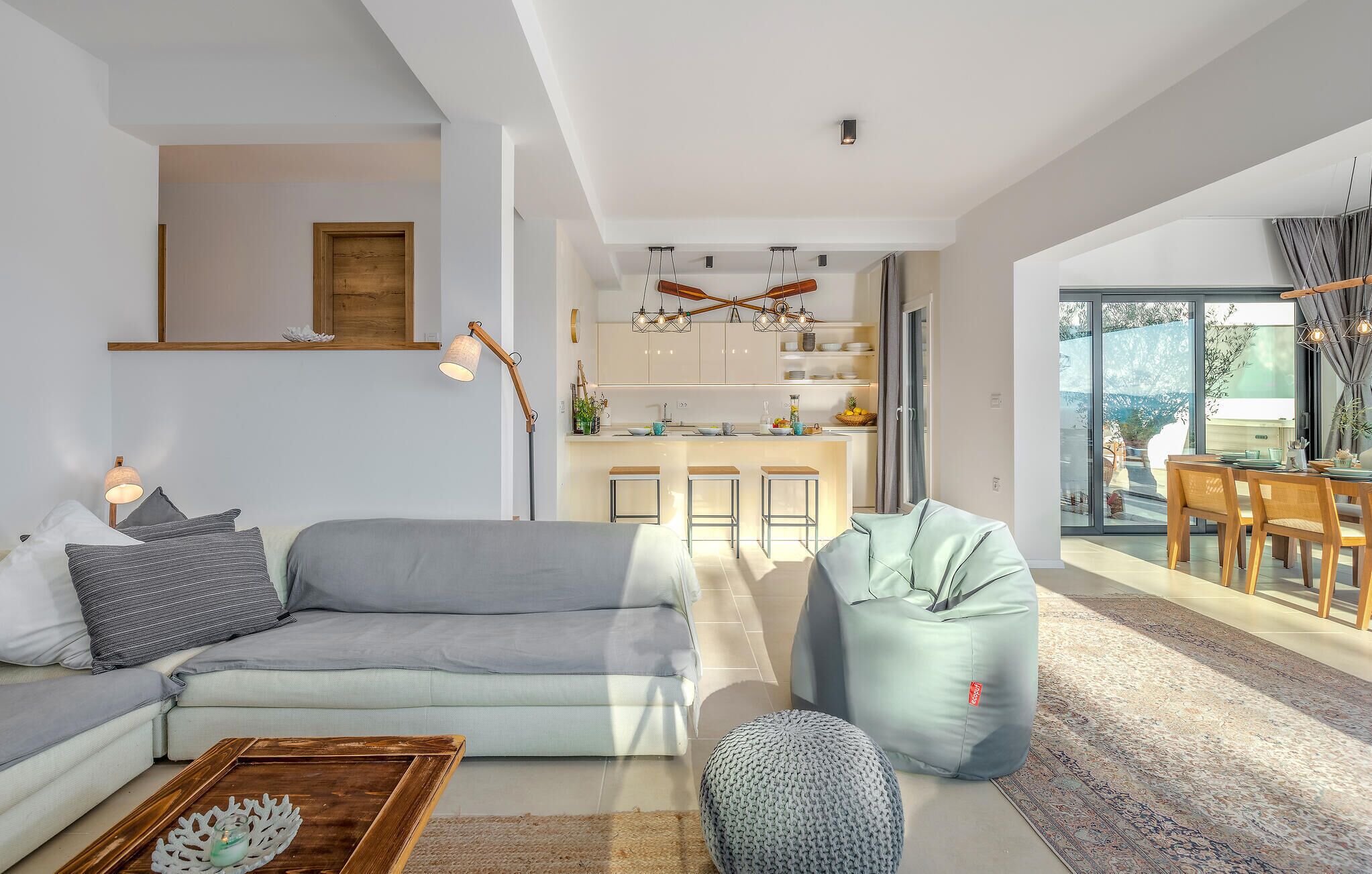 Een moderne woonkamer met een grijze bank, een houten salontafel en een relaxfauteuil. Door een open deur is een keuken met witte kastjes en een eetgedeelte met een tafel en stoelen zichtbaar.