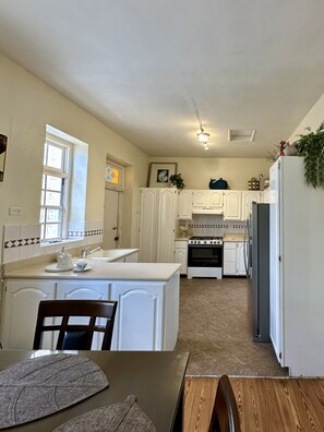 A kitchen with white cabinets, a stove, and a refrigerator. There is a window with a stained glass design and a plant on the counter. - Beach House Near Carlisle Bay, Walk to Beach, Historic Charm, Sleeps 4-6 (Bridgetown)