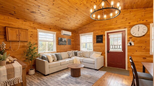 Een gezellig interieur van een chalet met houten plafond, een groot raam, een bank, een eettafel en een klok aan de muur.