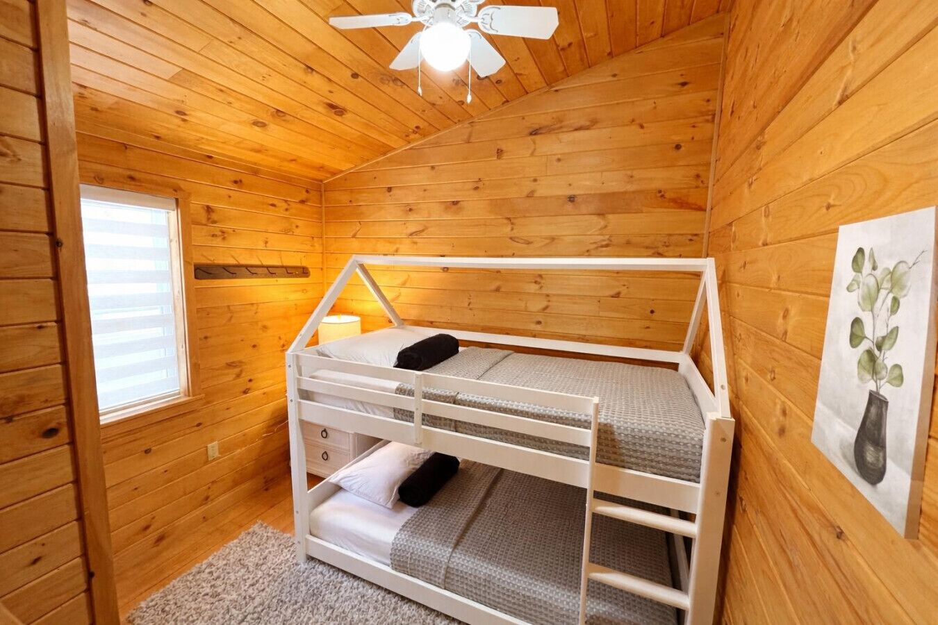Habitación de cabaña de madera con una cama litera, ventilador de techo, ventana con persianas y un cuadro en la pared.