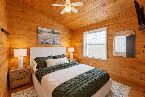 A wooden-paneled bedroom with a bed, two bedside tables, a ceiling fan, a window, and a painting on the wall.