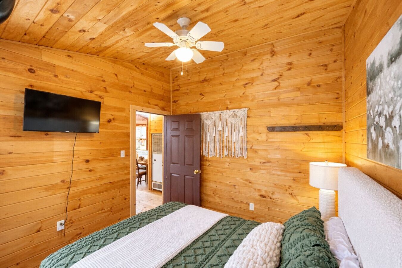 A wooden cabin bedroom with a bed, a ceiling fan, a TV, a door, and a wall-mounted lamp.
