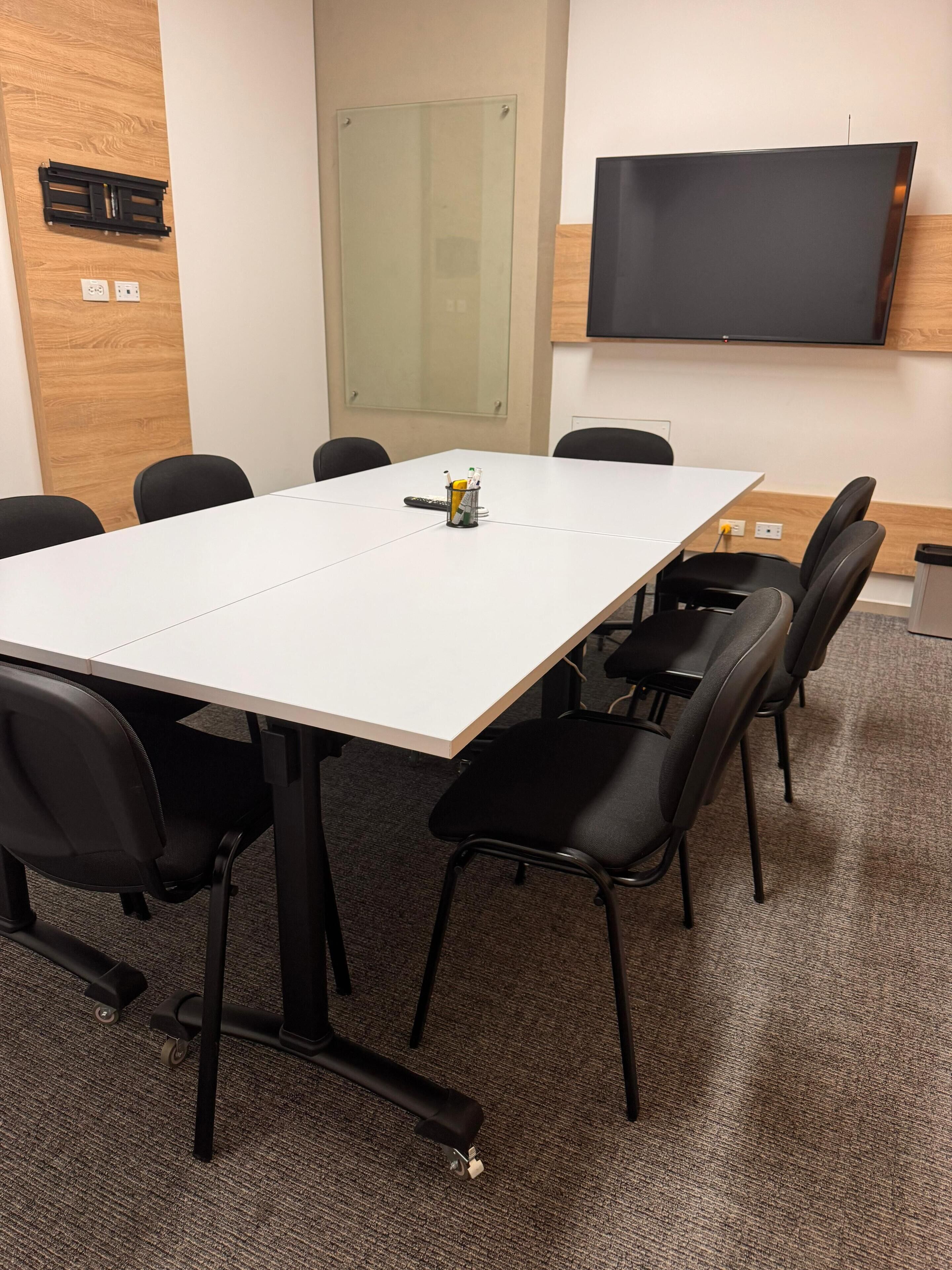 Una sala de conferencias con una mesa blanca, sillas negras, un televisor montado y una puerta de cristal.