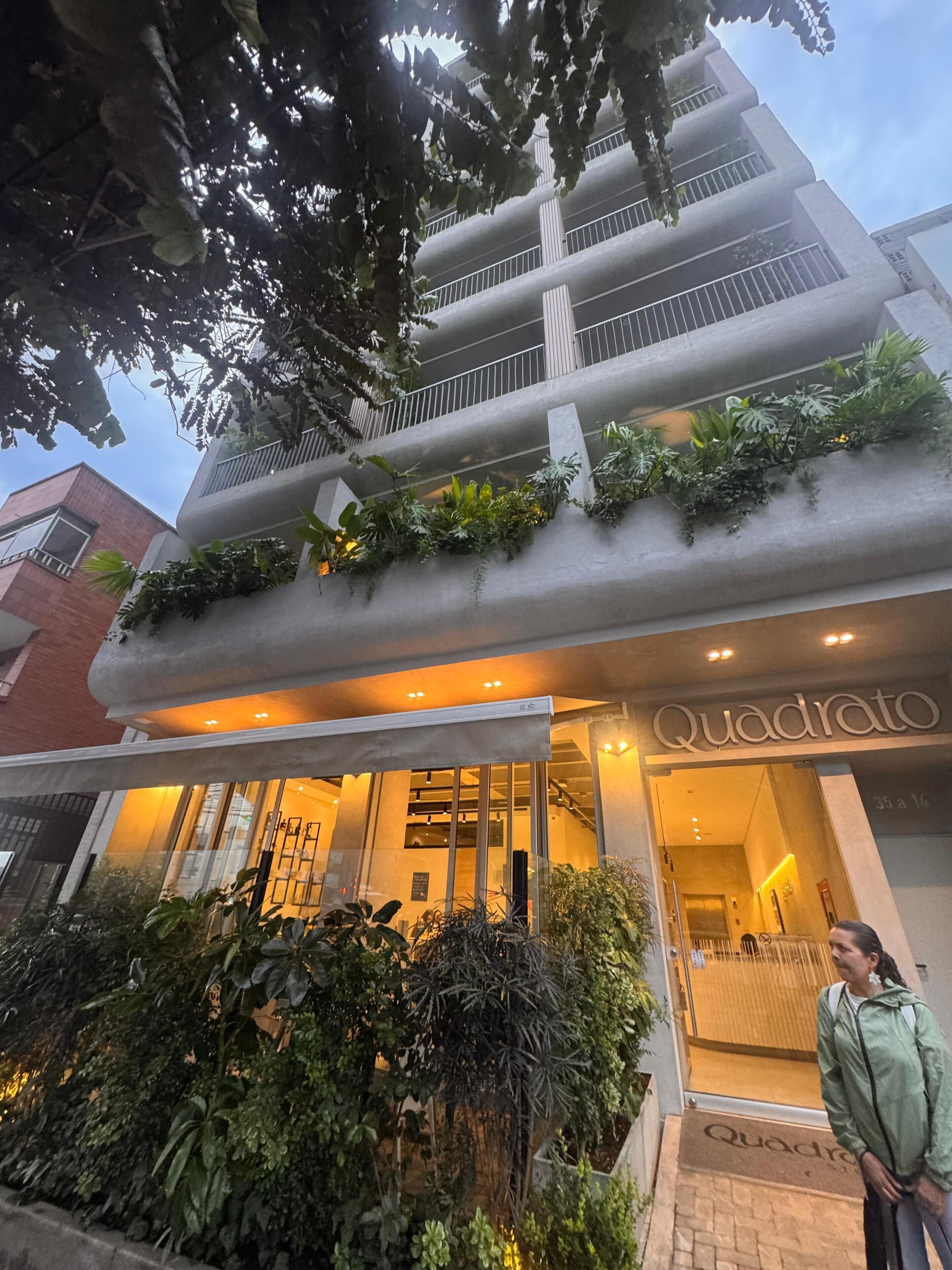 A multi-story building with balconies, a sign reading "Quadrato," and a well-maintained garden area.
